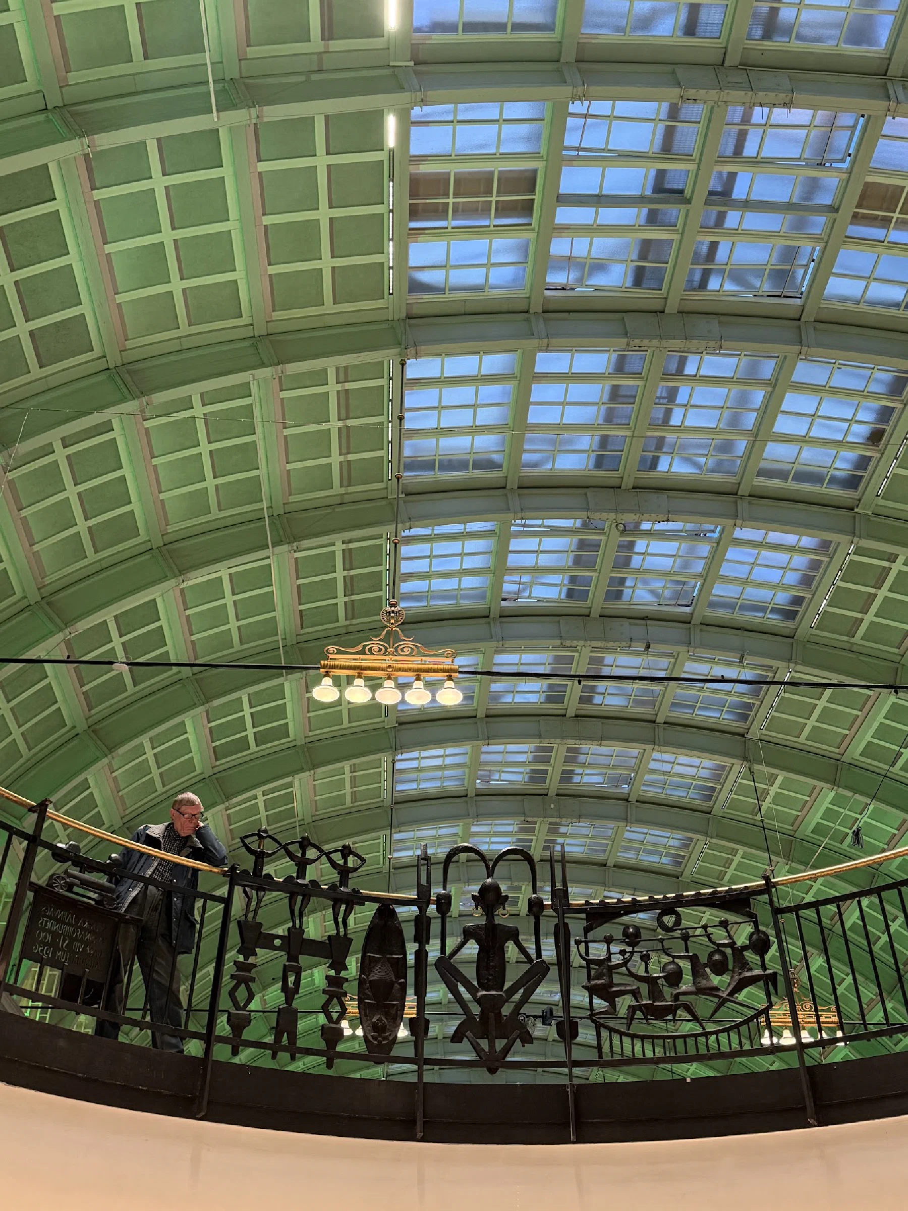 A pale green vaulted glass roof above a balcony lined with metal figures